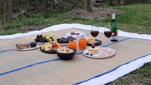 Un montaje para picnic con comida y bebidas sobre una manta, en un entorno natural con árboles y colinas al fondo.