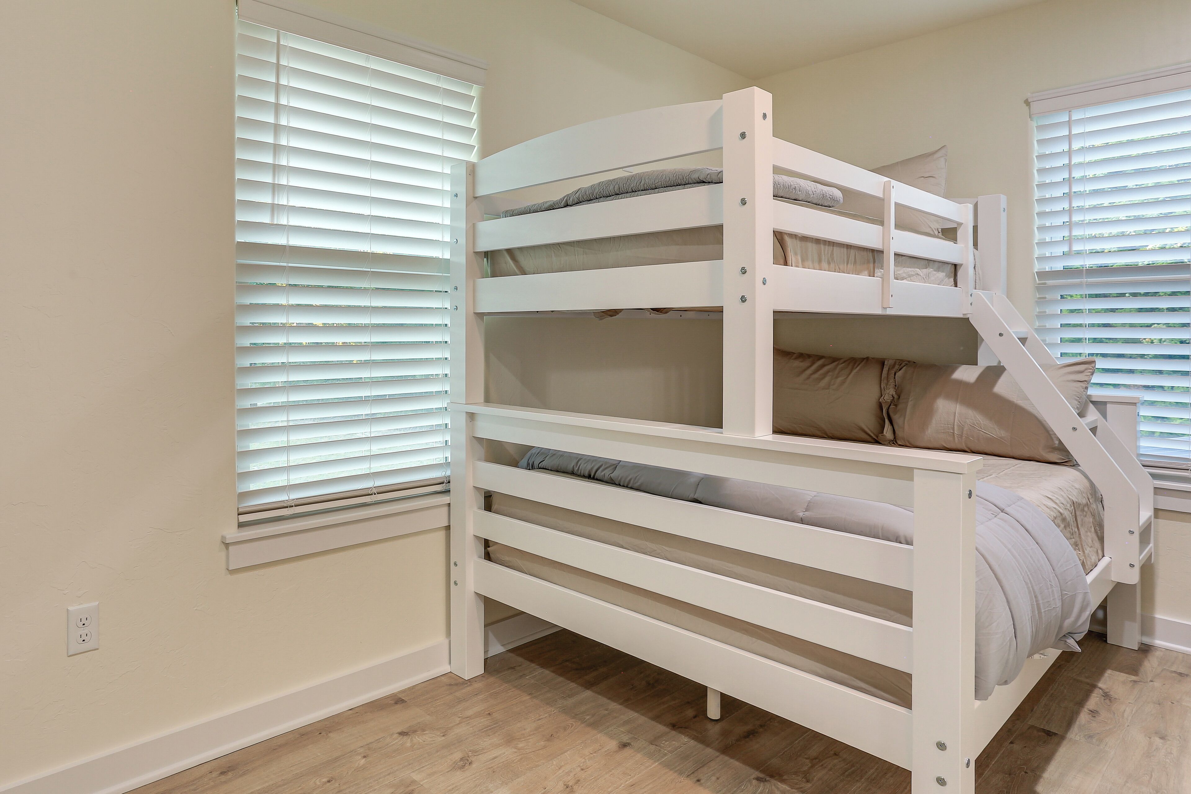 A bunk bed with a wooden frame and beige bedding, positioned in a room with wooden flooring and white walls.