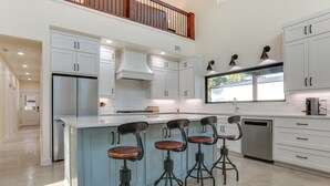 Eine moderne Küche mit weißen Schränken, einem Edelstahlkühlschrank, einer großen Kochinsel mit Barhocker und einem Fenster, das natürliches Licht hereinlässt.