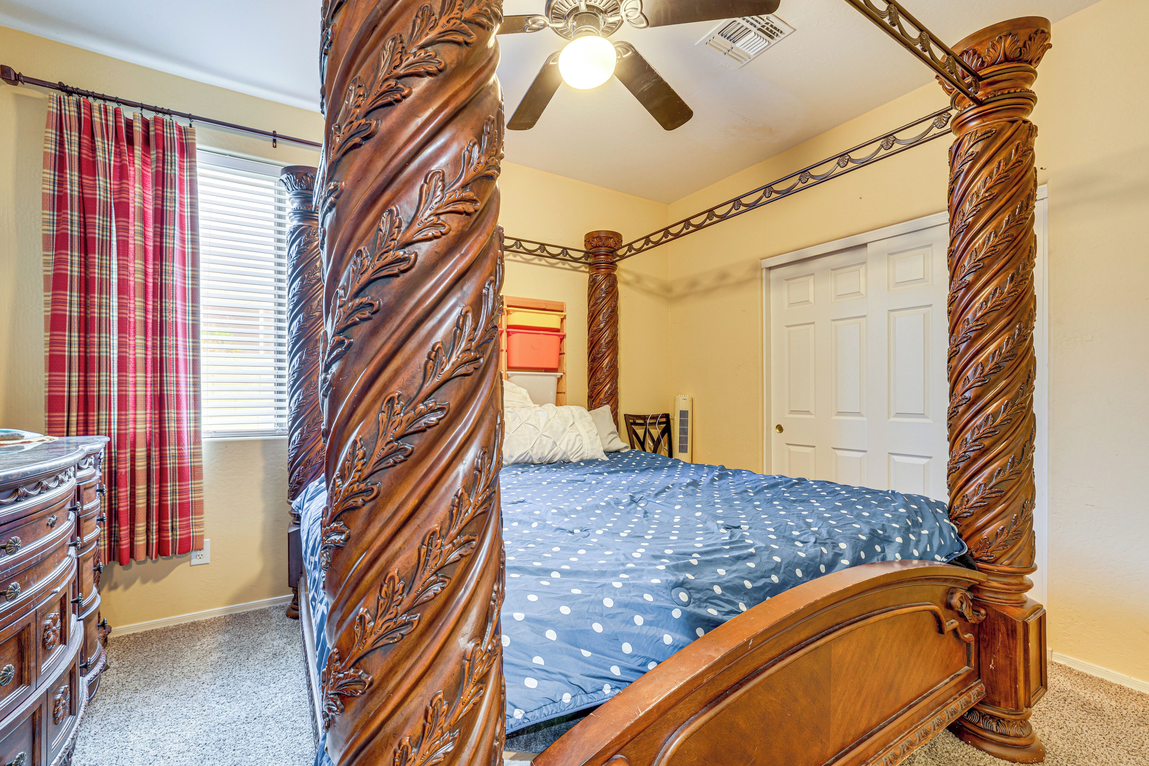 A four-poster bed with a wooden headboard and footboard, a ceiling fan, a dresser with drawers, and a window with plaid curtains.