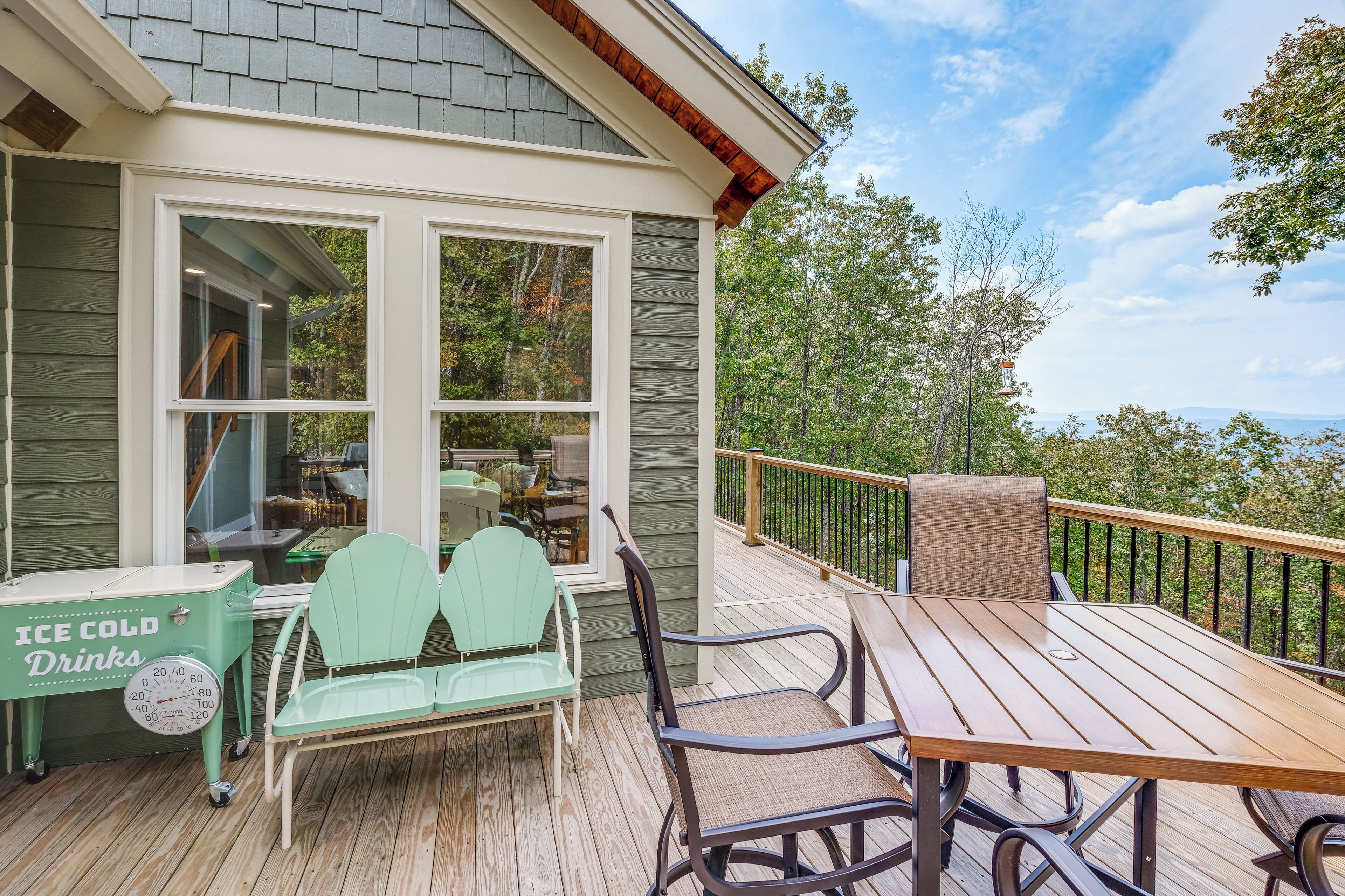 A wooden deck with outdoor furniture, a view of trees and mountains, and a house with a large window.