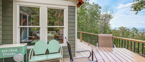 A wooden deck with outdoor furniture, a view of trees and mountains, and a house with a large window.
