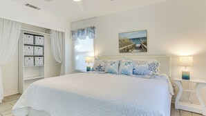 Un dormitorio con una cama grande, una mesita de noche con lámpara, una ventana con cortinas, un cuadro en la pared y un mueble con cajones.