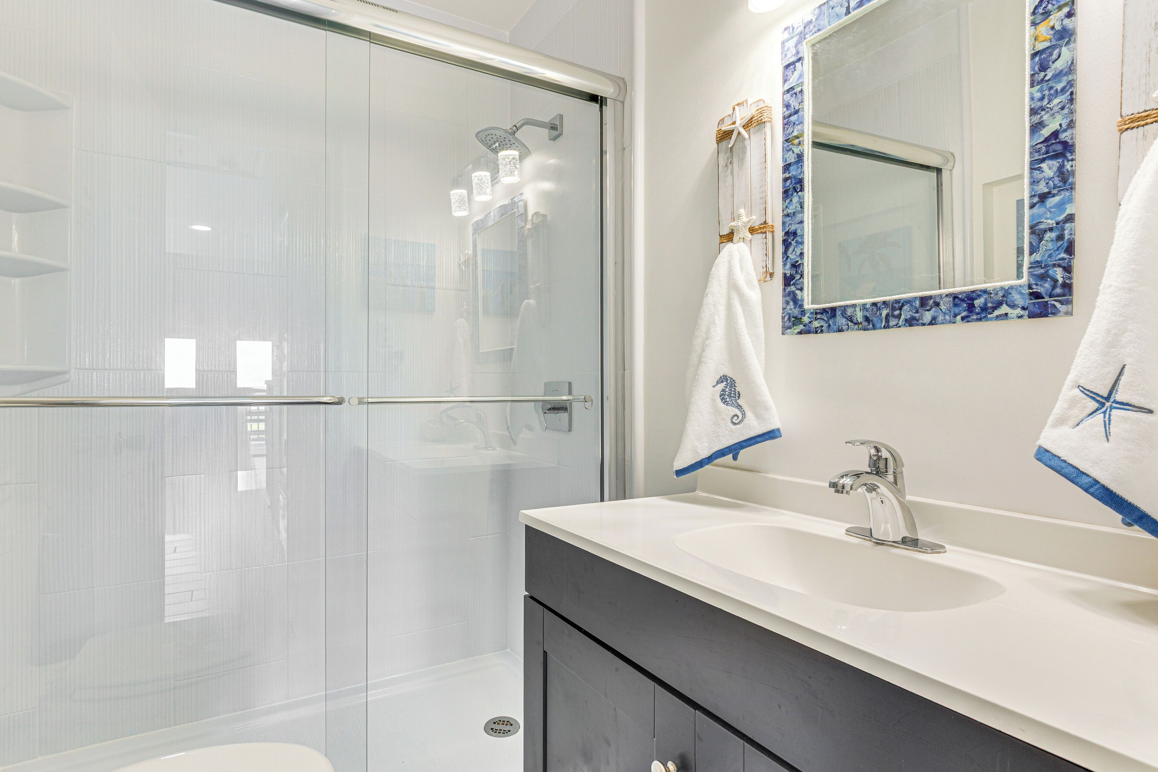 Een moderne badkamer met een glazen douchecabine, een witte wastafel met een donkere badkamerkast en een spiegel met een blauw-wit patroon.