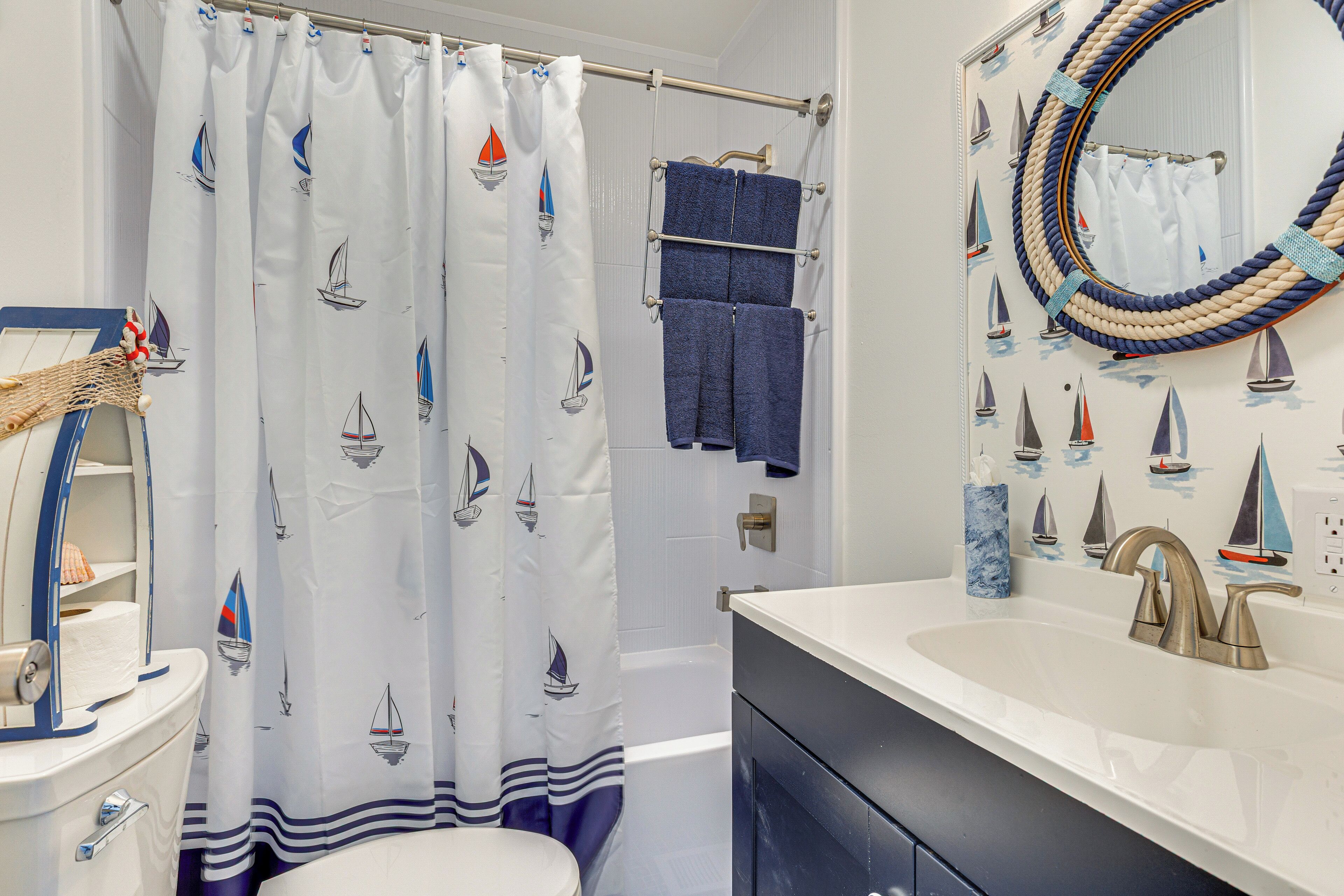 A bathroom with a nautical theme, featuring a shower curtain with sailboat designs, a towel rack with blue towels, and a mirror with a rope frame.