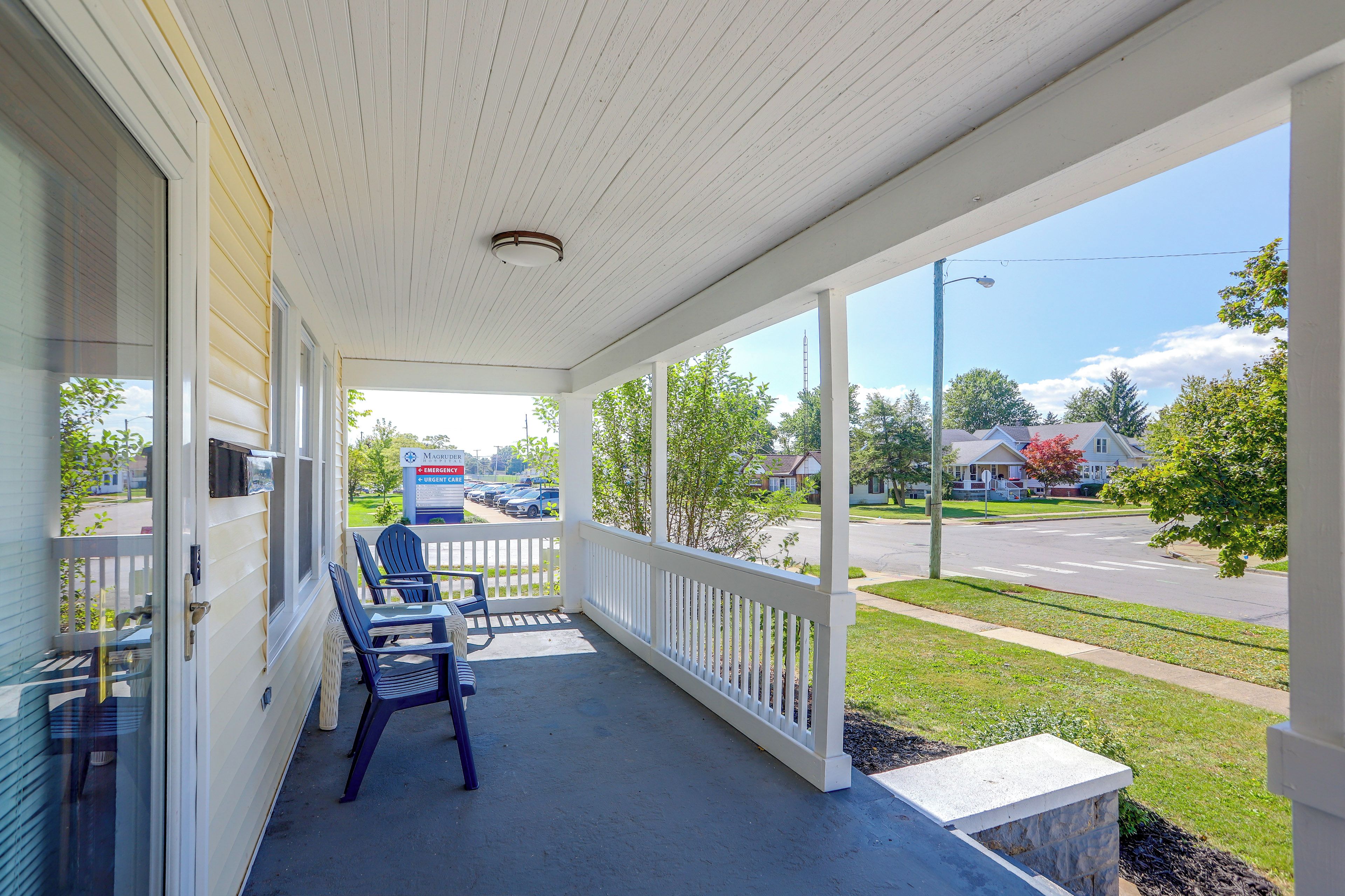 A covered porch with two chairs and a table, overlooking a street with parked cars and houses.