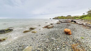 A pebbled beach with a wooden stump, calm sea, and a distant building on a grassy hill. - Fish & Hike: Quiet Smith River Cabin Near Ocean! (Smith River)