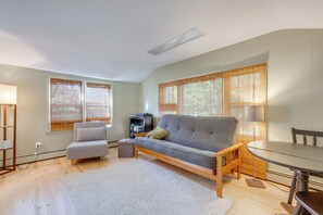 A modern living room with a grey sofa, a small table, and a chair. There are large windows with blinds, a lamp, and a bookshelf. - Home w/ Private Yard in Rocky Point (Rocky Point)