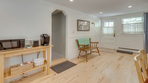 A clean, well-lit room with a wooden floor, a table with a microwave, a coffee maker, and a wicker basket. There is a chair with a green towel, a door, and a window with curtains. - Home w/ Private Yard in Rocky Point (Rocky Point)