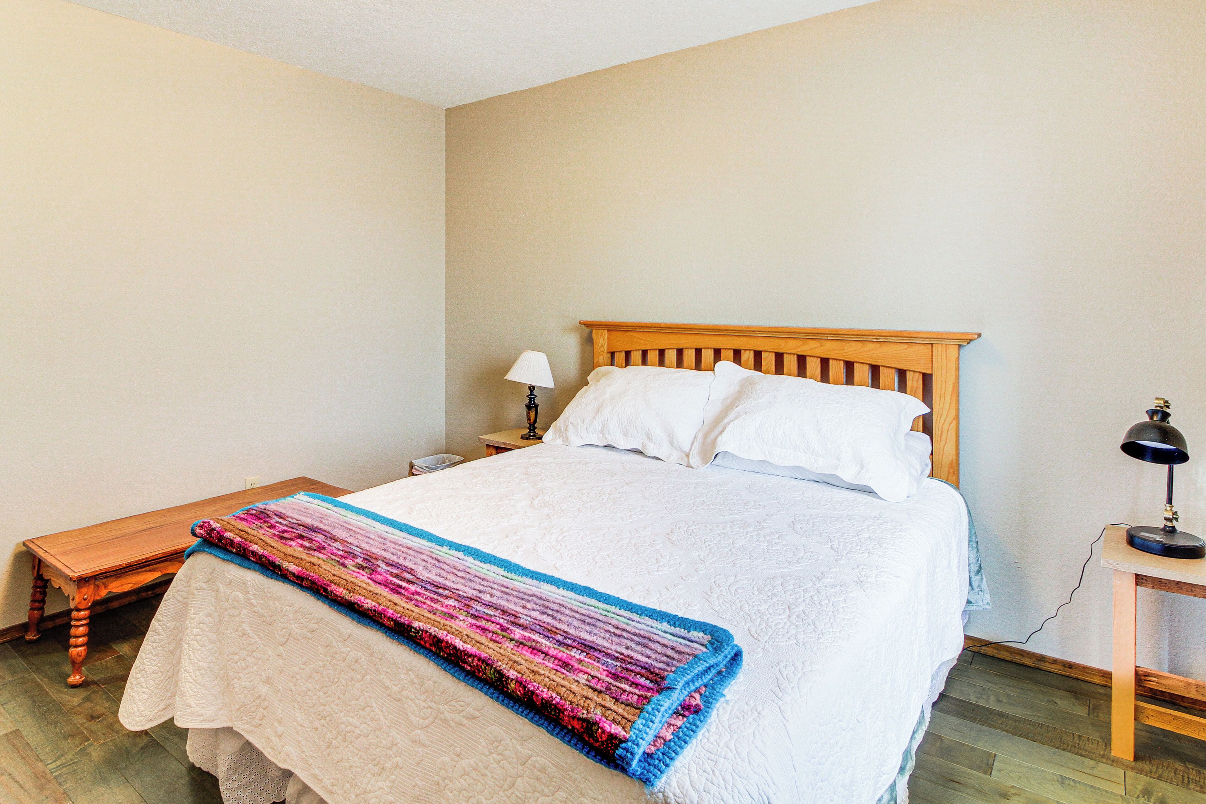 A bedroom with a wooden bed, a bedside table with a lamp, a small wooden bench, and a colorful blanket.