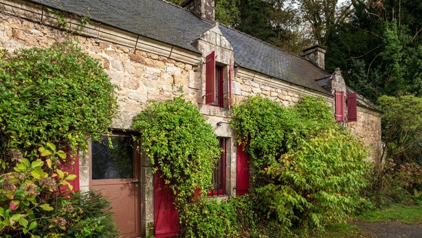 Ein Gebäude aus Stein mit roten Fensterläden, umgeben von viel Grün.