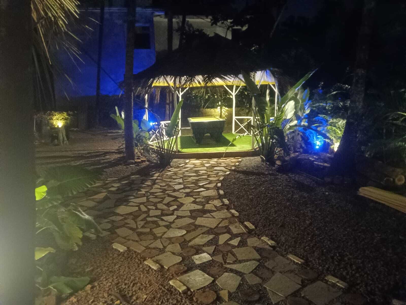A stone pathway leading to a gazebo with a thatched roof, surrounded by tropical plants and illuminated by blue lighting.