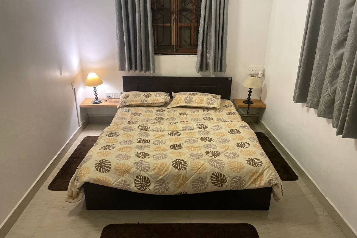 Una cama cuidadosamente tendida con una colcha de diseño de hojas, dos mesitas de noche con lámparas, una ventana con cortinas y una alfombra marrón en el suelo.