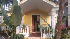 A yellow building with a white porch, a stone staircase, and tropical plants.