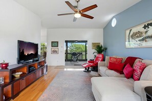 Una sala de estar con televisor, sofá y cojines rojos, ventilador de techo y vista al exterior.