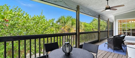 Een overdekt terras buiten met een ronde tafel en stoelen, een plafondventilator en uitzicht op weelderige begroeiing.
