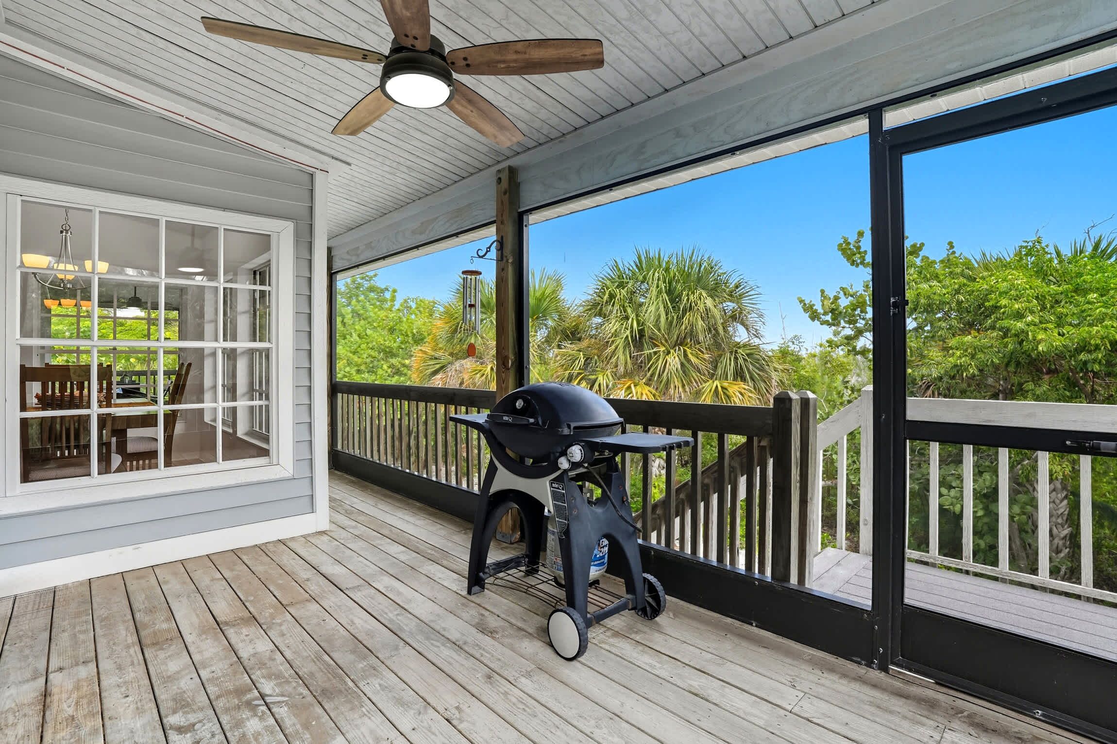 A covered outdoor deck with a grill, a dining area with a table and chairs, and a view of greenery.