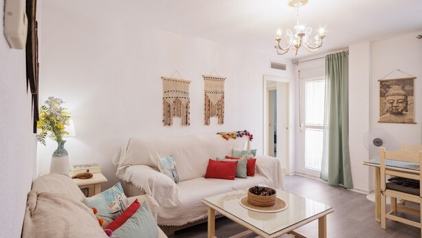 A living room with a white sofa, a small table, a fan, and a Buddha painting on the wall. - Stunning Views of Seville (San Juan de Aznalfarache)