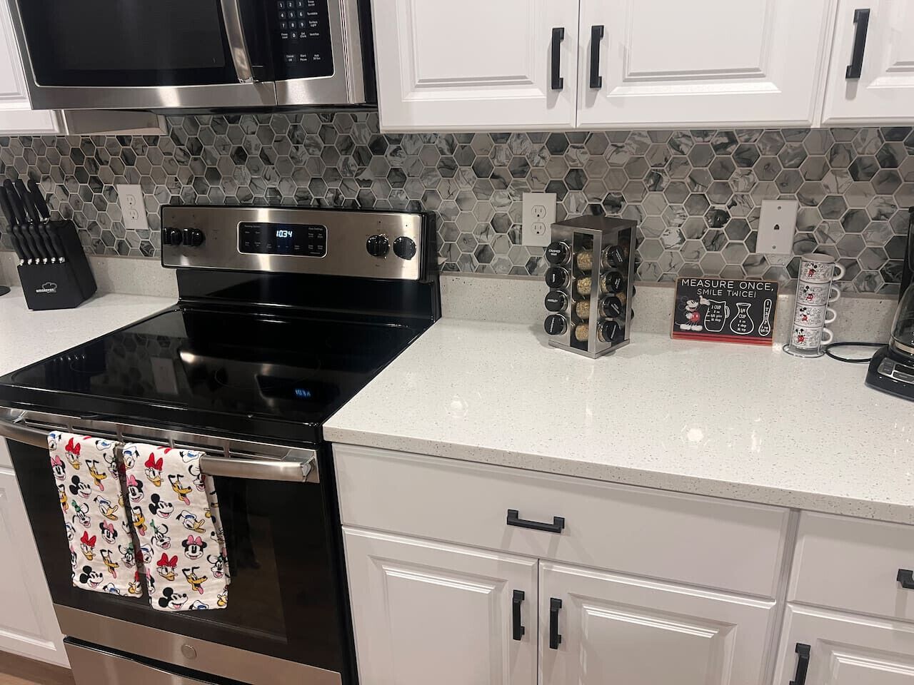 A kitchen with a black stove, white cabinets, and a microwave above it. There are measuring cups on the counter and a sign that says "MEASURE ONCE, SMILE TWICE."