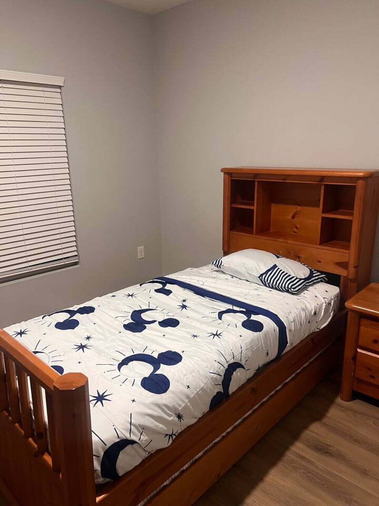 A single bed with a wooden headboard and footboard, a bedside table, and a window with blinds.
