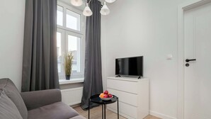 A modern living room with a grey sofa, a small black table with fruit, a TV on a white cabinet, and a door with a handle. - Cozy Apartment | Close to Manufaktura and Piotrkowska Street (Łódź)