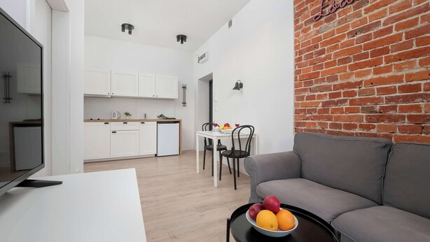 A modern living room with a grey sofa, a small table with fruit, and a brick wall with the word "Lodge" on it. - Cozy Apartment | Close to Manufaktura and Piotrkowska Street (Łódź)
