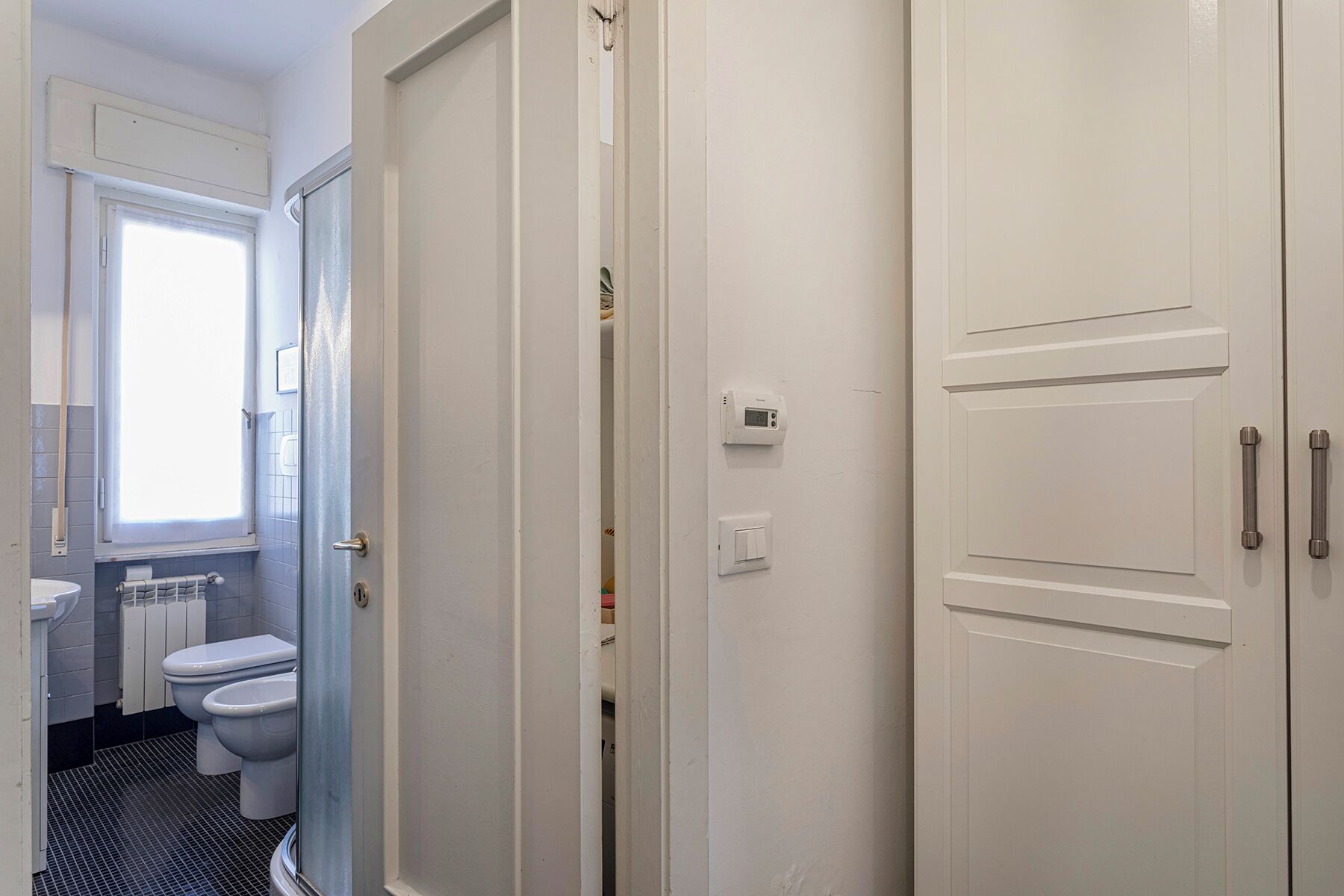 A bathroom with two toilets, a window, a radiator, and a white door with a handle.