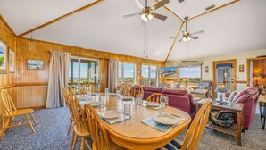 Ein geräumiger Raum mit einem Holzespeisetisch, gedeckt für vier Personen, umgeben von Holzstühlen. Zwei Deckenventilatoren, ein großes Fenster mit Blick auf den Strand und eine gemütliche Sitzecke mit einem roten Sofa und Sesseln.