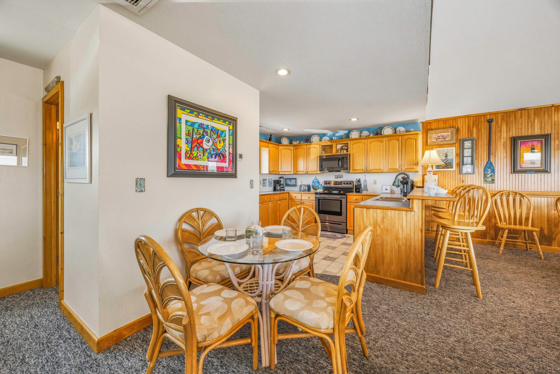 A compact kitchen with wooden cabinets, a microwave, and a stove. There is a dining area with a glass table and wicker chairs.