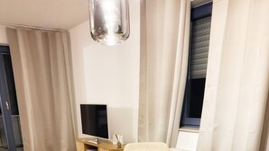 Uma sala de jantar com uma mesa redonda de madeira, cadeiras brancas, uma televisão pequena sobre um suporte de madeira e uma janela com cortinas.