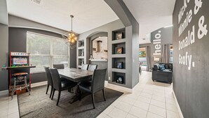 A modern kitchen with a dining area, featuring a built-in arcade game, a chandelier, and a wall with a playful message. - Adventure Awaits Manor (Kissimmee)