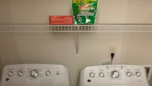 Dos lavadoras de carga frontal blancas, una al lado de la otra, con una caja de detergente verde en un estante superior.