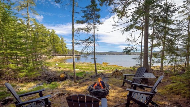 Leirintäalue järven rannalla, jossa on kolme Adirondack-tyylistä tuolia, nuotiopaikka sekä näkymä järvelle ja kaukaisiin vuorille.