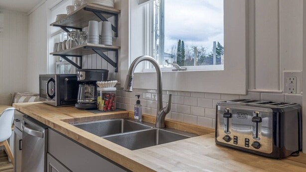 A kitchen with a double sink, a coffee maker, a toaster, and shelves with dishes and glasses. - Renovated 3BR Home in Downtown SWH [Mariners Rest] (Southwest Harbor)