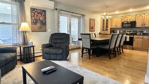 Een moderne woonkamer met een donkere bank, een salontafel en een eethoek met tafel en stoelen. Achtergrond is een keuken met houten kastjes en een magnetron.