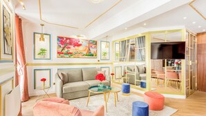 A modern living room with a sofa, coffee table, and ottoman, a painting on the wall, and a TV in a wooden cabinet. - Merveil Luxury Suite - Aboukir VII (Paris)