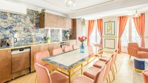 A modern kitchen with a marble countertop, a built-in dishwasher, and a dining area with pink chairs and a glass table. - Merveil Luxury Suite - Aboukir VII (Paris)