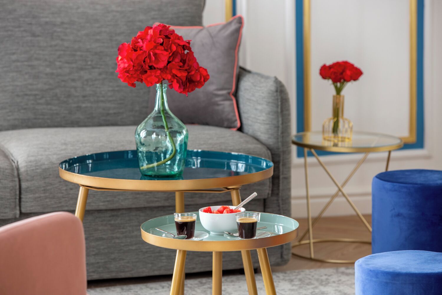 Een moderne woonkamer met een grijze bank, een rond glazen bijzettafeltje met een vaas rode bloemen, een schaal fruit en twee kleine glaasjes donker drankje.