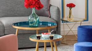 A modern living room with a grey sofa, a round glass coffee table with a vase of red flowers, a bowl of fruit, and two small glasses of a dark beverage. - Merveil Luxury Suite - Aboukir VII (Paris)
