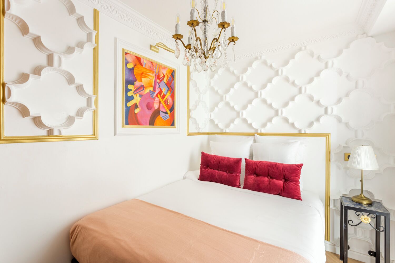 A bedroom with a white bed, red pillows, a chandelier, and a painting on the wall.