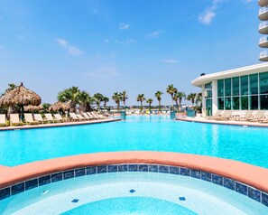 A swimming pool with lounge chairs and palm trees, surrounded by a building with large windows. - Caribe 4BR, Panoramic Gulf Views, Lazy River, Hot Tub, 2 Min To Beach, Sleeps 12 (Orange Beach)