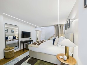 A modern hotel room with a large bed, a flat-screen TV, a bedside table with a lamp, and a large mirror.