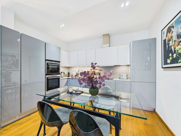 A modern kitchen with a glass dining table, two chairs, and a vase of flowers.