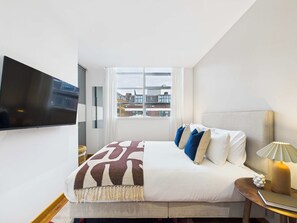 Une chambre d’hôtel avec un lit, un téléviseur fixé au mur, une fenêtre donnant sur des immeubles et une table de chevet avec une lampe.