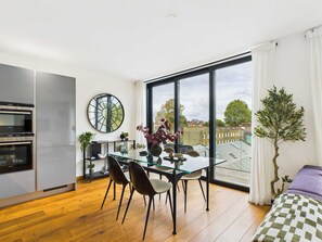 Une cuisine moderne dotée d’appareils électroménagers intégrés, un coin repas avec une table et des chaises en verre, et une grande fenêtre offrant une vue sur la ville.