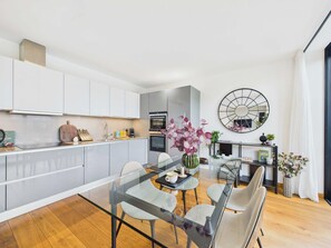 Une cuisine moderne avec des meubles blancs, un four encastré et un coin repas comprenant une table et des chaises en verre.