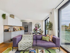 Un salon moderne avec un canapé violet, une table basse en verre et une vue sur la ville grâce à de grandes fenêtres.