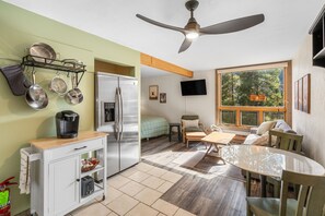 A compact kitchen with a refrigerator, coffee maker, and hanging pots. There's a dining area with a table and chairs, a sofa, and a TV mounted on the wall.