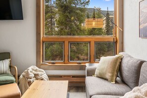 A cozy room with a large window, a sofa, a chair, and a wooden table. - Big Sky Resort Studio | Walk to the Shuttle (Big Sky)