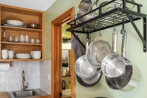 Uma cozinha com uma estante de madeira com pratos e copos, uma grade de metal com panelas e frigideiras, e uma pia com torneira.